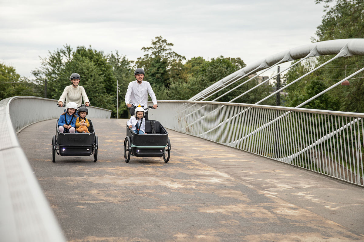 Two families riding Black Iron Horse cargo trikes across a footbridge, children in the front boxes