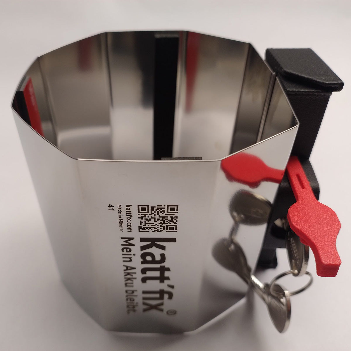 Kattfix stainless-steel battery lock band from above, showing the branding, QR code and red fitting keys